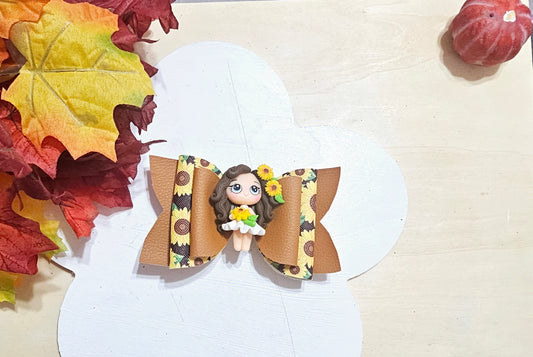 Handmade sunflower-themed hair bow with brown and yellow layers and a decorative girl centerpiece, displayed with autumn leaves.
