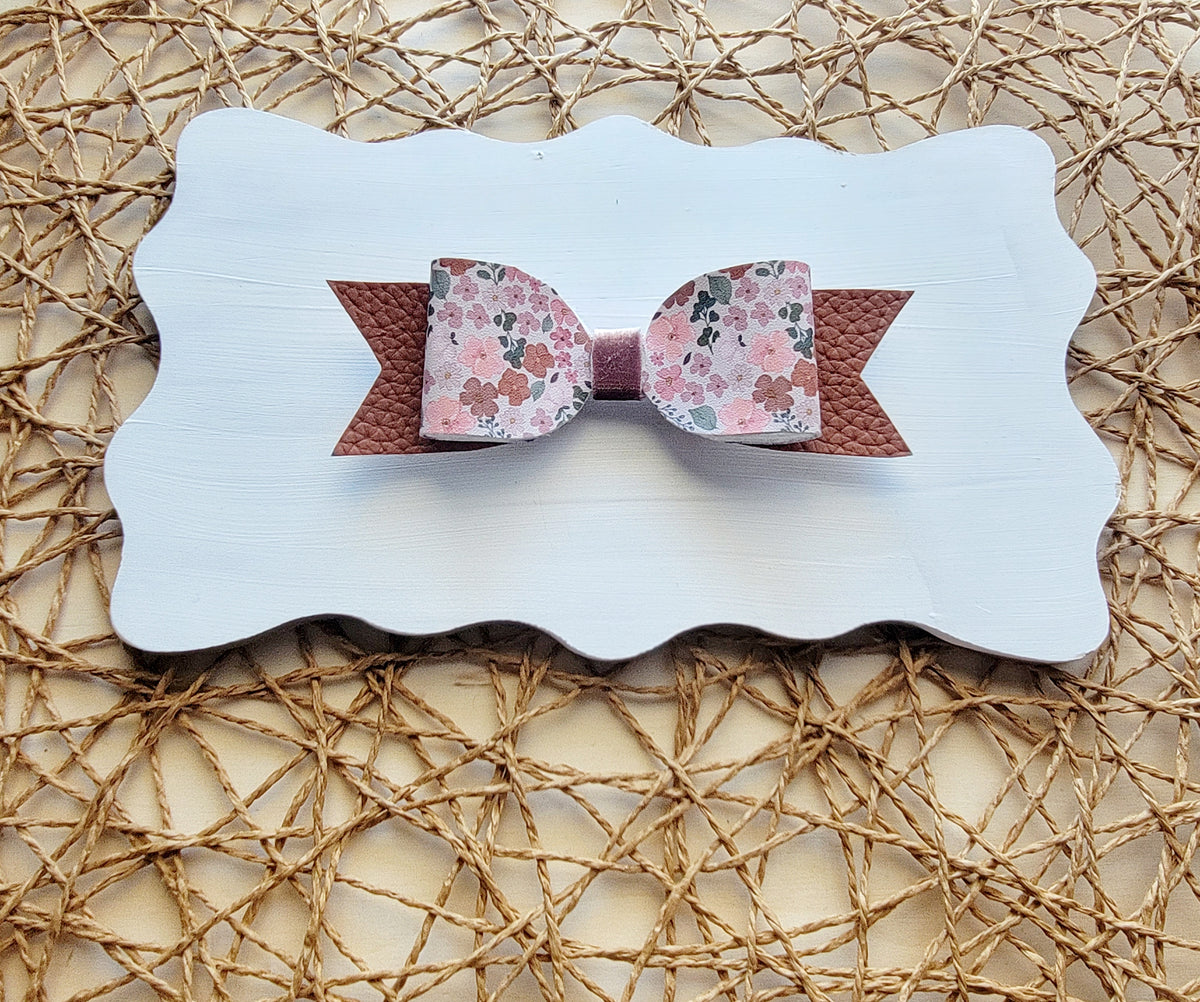 Handmade pink floral hair bow with a soft pink center and rose-gold glitter ribbon tails, displayed on a white board.